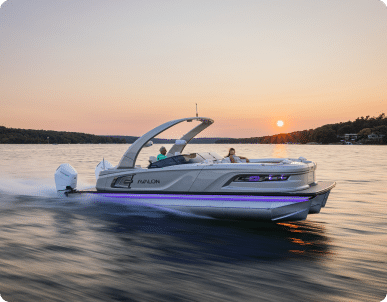 A sleek motorboat with passengers cruises on a calm body of water at sunset. The boat's lights are illuminated, casting a subtle glow. The sky is serene, with the sun partially set, and the shore is visible in the background, resembling the tranquil scenes enjoyed by luxury pontoon boats.