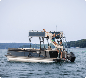 A Pontoon boat floats on a lake. Several people are on the boat, with one person sliding down a waterslide attached to the upper deck. The lake is surrounded by distant, tree-covered hills under a clear sky. Imagine enjoying this scene every weekend with pontoon boats for sale now!