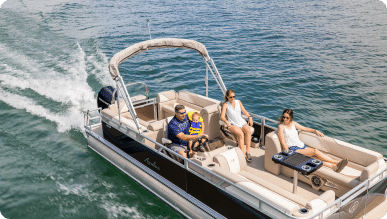 A family enjoys a sunny day on a pontoon boat in open water. The father steers while the child, wearing a life jacket, sits on his lap. The mother and another woman relax on the seating area, soaking up the sun as the boat creates a small wake behind it—a perfect moment to consider one of these pontoon boats for sale.