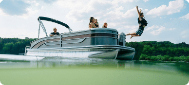 A group of people enjoying a sunny day on a pontoon boat for sale. One person is captured mid-air as they jump off the boat into the water. The boat is on a calm lake with green hills in the background.