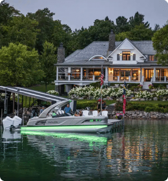 A sleek, modern pontoon boat with green underlighting is docked in front of an elegant lakeside house in the evening. The house has large windows and a wraparound porch adorned with American flags and lush greenery. Explore the finest pontoon boats for sale and elevate your lake experience today.