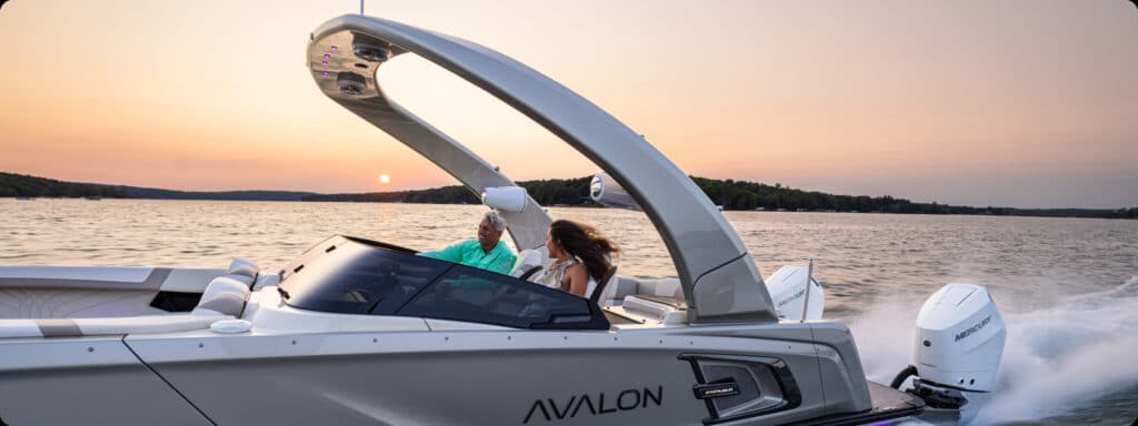 A sleek white Avalon motorboat, reminiscent of the new pontoon boats for sale, speeds across a calm lake during sunset. Two people are seated in the boat, enjoying the ride. The water reflects the colors of the sunset, and forested hills line the horizon in the background.