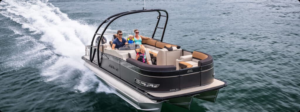 A family of four, including two adults and two children, are enjoying a ride on one of the new pontoon boats for sale in open water. The boat is moving at a high speed, creating a wake behind it. The day is clear, and everyone on the boat is wearing life jackets.