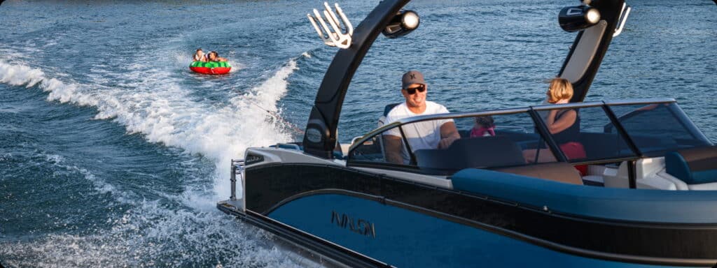 A man and a woman are riding in a sleek blue motorboat, navigating through a lake. Behind the boat, two children in a green and red inflatable tube are being towed, bouncing on the water's surface and enjoying the thrill of the ride. Nearby, new pontoon boats for sale glisten under the sun's rays.