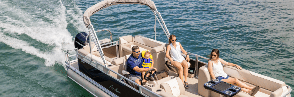 Three adults and a child are enjoying a sunny day on one of the small pontoon boats. The man at the wheel is steering, while the child, wearing a yellow life jacket, sits on his lap. Two women are seated comfortably on the boat's cushioned seats as it cruises on a calm, blue lake.