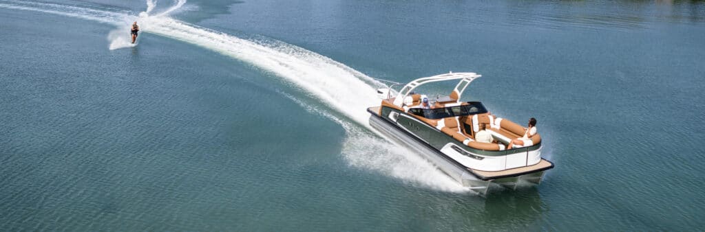 A spacious motorboat with several people on board cruises on a calm lake, pulling a waterskier behind it. The boat leaves a trail of white wake in the water. Nearby, pontoons for sale dot the shore, adding to the relaxed atmosphere as the skier glides smoothly on the water.