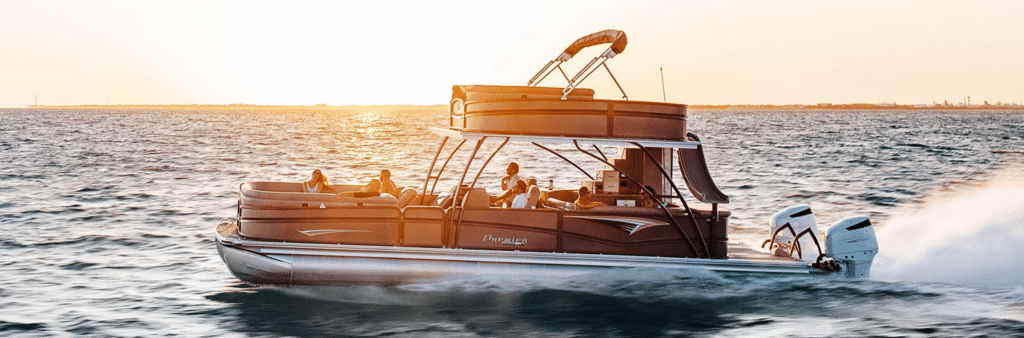 A pontoon boat with several people on board is cruising on a body of water at sunset. The boat, one of the best pontoon boats around, has two outboard motors and features a canopy. The water is slightly choppy, and the sun is low in the sky, casting a warm glow over the scene.