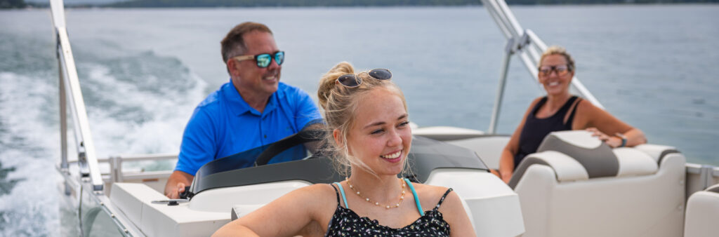 Three people enjoy a boat ride on a lake. A young woman with blonde hair and sunglasses rests at the front, smiling. The man in a blue shirt and sunglasses pilots the boat, while the woman behind him, wearing a black top and sunglasses, smiles at the camera amidst scenic pontoons near me.