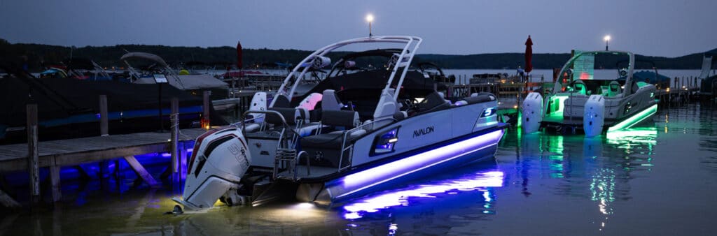 A sleek motorboat with vibrant purple and green underwater lights is docked at a pier in the evening. Nearby pontoons near me enhance the scenery, while other boats and a serene lake in the background, along with subtle lighting and a twilight sky, create a calm and inviting atmosphere.
