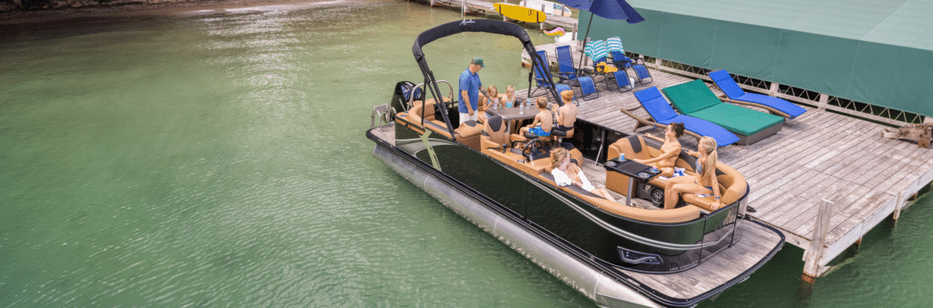 A boat filled with people enjoying a day on the water is docked beside a large green-covered pier. The pier has lounge chairs, towels, and a closed green umbrella. The surrounding water is calm and greenish, matching the peaceful ambiance of the scene—perfect for those searching pontoons for sale near me.