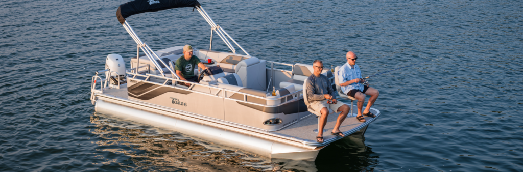 Three people are fishing on a sunny day aboard a pontoon boat they found by searching "pontoons near me." The boat floats on calm water, with one person steering and two sitting with fishing rods. It features a shade canopy, cushioned seats, and various fishing gear.