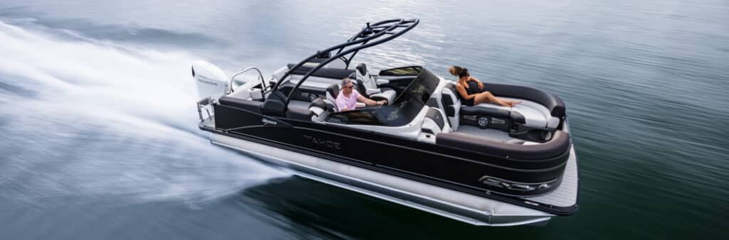 A sleek black pontoon boat speeds across the water with two people aboard. One person stands at the helm steering while the other lounges on a cushioned seat, relaxing under the sun. The sturdy pontoons create a trail of white waves behind it, cutting through the calm water.