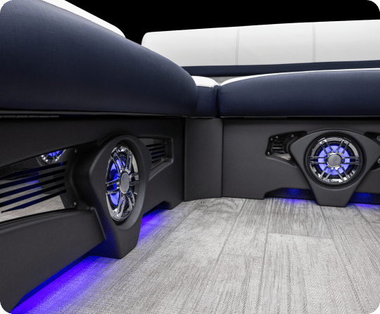 Close-up view of a boat's seating area, highlighting built-in speakers and ambient blue LED lighting underneath the seats. The floor is made of a grey wood-like material. The boat's seating is upholstered in dark and light colors, perfect for those browsing pontoons for sale.