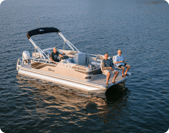 A pontoon boat floats on a body of water with three people onboard. Two people sit on the deck's edge with feet dangling over the pontoons, while the third person sits inside under the boat's canopy. The water is calm, reflecting the boat and the sunset sky.
