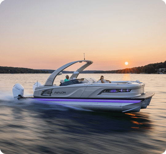 A luxury pontoon boat with "Avalon" branding glides across a calm lake at sunset, featuring sleek design elements and an arching roof structure. The sun is low on the horizon, casting an orange glow on the pontoons and water.