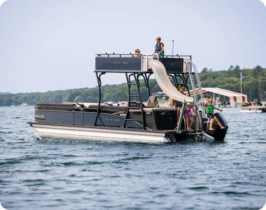 A pontoon boat on a lake with several people onboard. Two children are at the top of a slide, attached to one of the pontoons, preparing to slide down while others watch from the lower deck. Trees are visible in the background, and the water is calm.