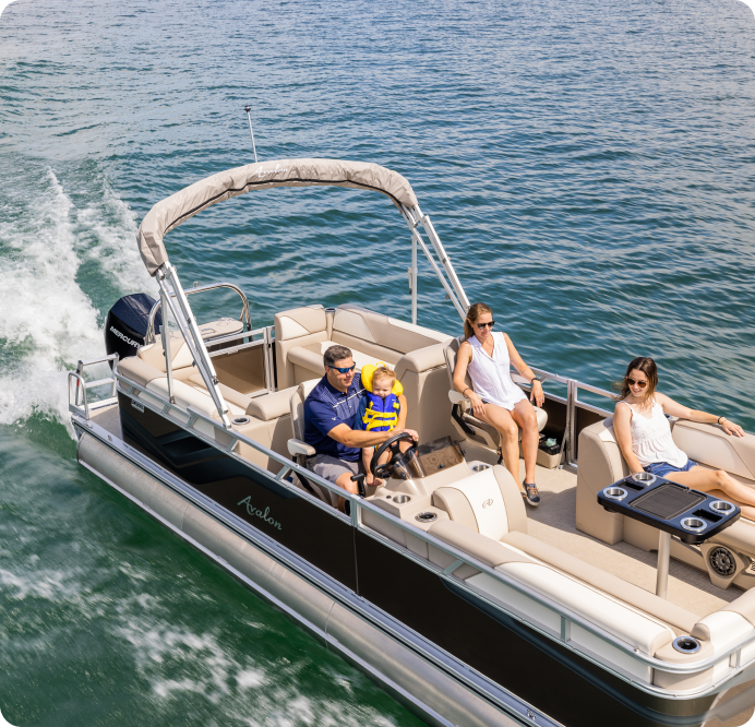 A family enjoys a ride on a pontoon boat on a sunny day. A man is steering the boat, holding a child wearing a yellow life jacket. Two women are seated, one in a recliner, relaxing and soaking up the sun as they glide swiftly over calm, blue water—easily found by searching for pontoons near me.