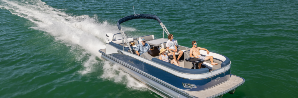 A pontoon boat speeds across the green water with five people on board. The boat has a blue canopy and sleek design, common among fishing pontoons. Two adults sit at the helm, while three others relax on the cushioned seats, enjoying the sunny day.