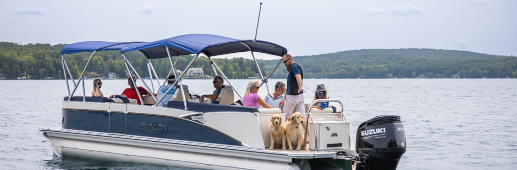 Several people and two dogs are enjoying time on a pontoon boat with a Suzuki engine on a lake. The boat, likely acquired from a local pontoons dealer, is anchored or moving slowly. Surrounded by trees and some houses in the background, the group sits under a shaded canopy, engaging in conversation.