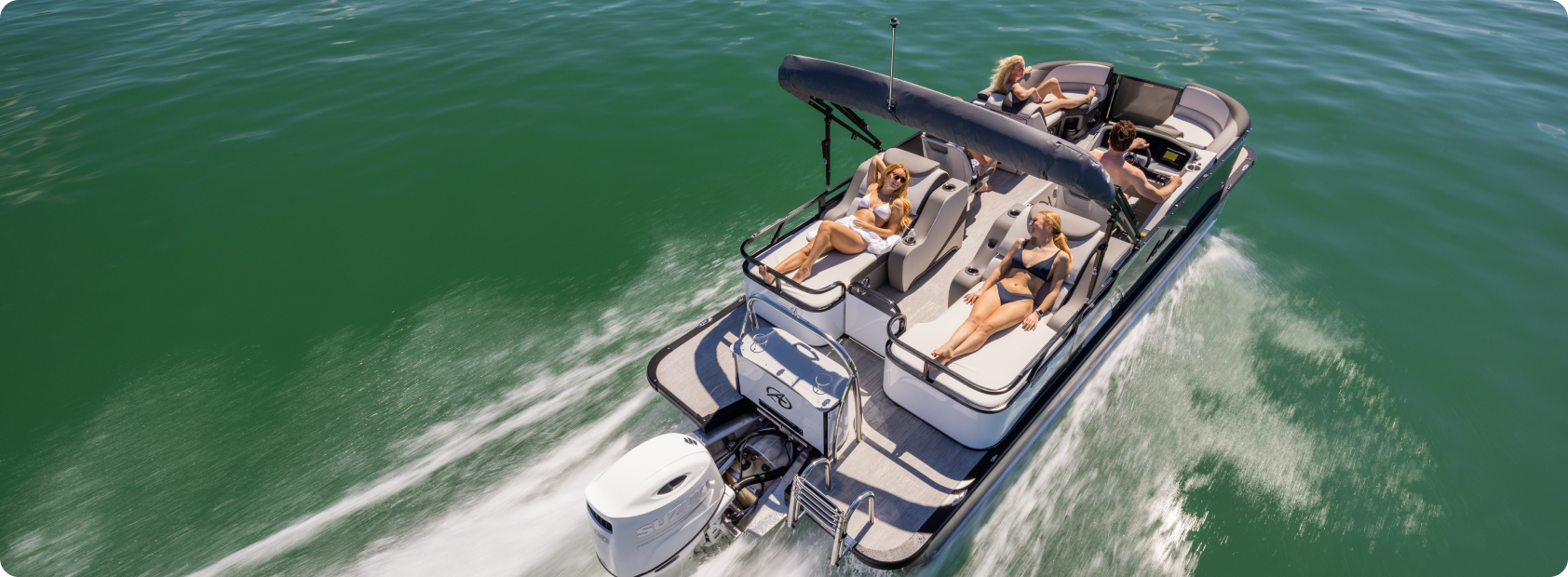 Aerial view of a motorboat cruising on clear green waters. The boat, resembling luxurious fishing pontoons, has a canopy and several passengers relaxing on deck chairs and in the lounge area. Two women sunbathe on the front deck while others sit and enjoy the ride. The boat's wake can be seen behind.