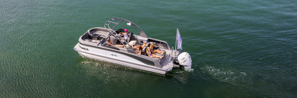 A small motorboat with four people lounging on cushioned seats and sunbathing is cruising on a calm, greenish body of water. The boat, which tempts anyone to consider buying pontoons for their own adventures, has a sleek design with a canopy frame and a visible outboard motor at the rear. The water appears still and serene.