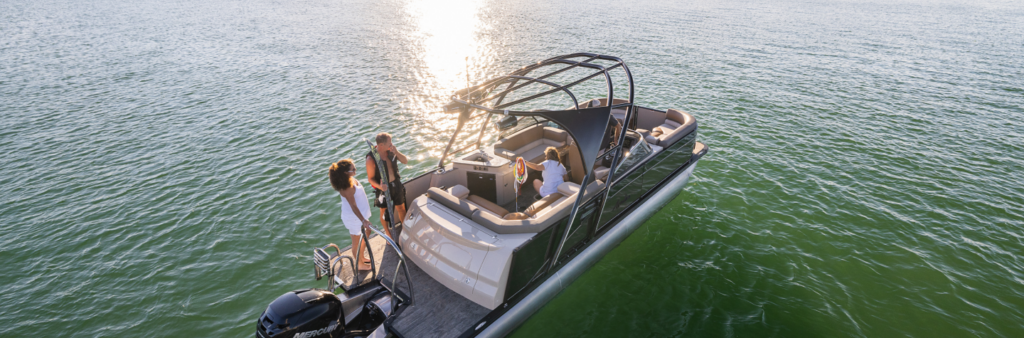 A group of people enjoying a day on a sleek, modern motorboat on a calm body of water during sunset. The boat has a canopy and appears spacious with comfortable seating, reminiscent of used pontoons updated for luxury. The sun creates a glistening reflection on the water's surface.