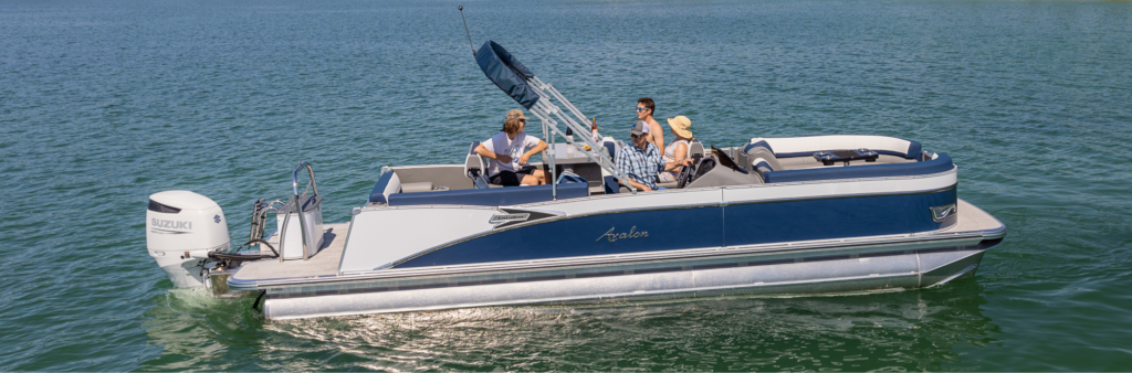 A group of people are on a blue pontoon boat named "Aquila" with a Suzuki outboard motor. The boat, equipped with various pontoon accessories, is on a calm body of water, and the sun is shining brightly.