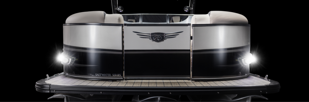 A front view of a sleek, modern pontoon boat with small pontoons and bright headlights shining against a black background. The boat has a metallic exterior with a logo and wing design on the front. The deck is wooden with a clean finish.