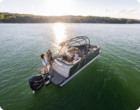 A boat with a dark hull navigates calm waters during a sunny day. Four people, two at the front and two at the rear, relax and enjoy the view. The water is green, and a forested shoreline is visible in the background, shimmering under the sunlight—an idyllic scene for those looking to buy pontoons.