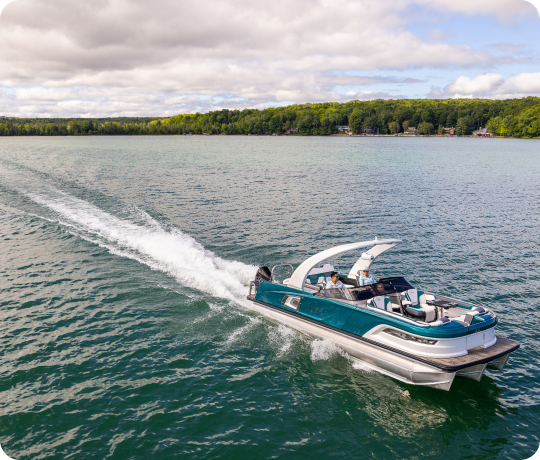 A sleek, modern teal boat speeds across a tranquil lake, leaving a white trail of wake behind it. Several people are onboard, enjoying the ride. The background features a lush, green tree-lined shoreline under a partly cloudy sky—a perfect scene showcasing our new pontoon boat sales.