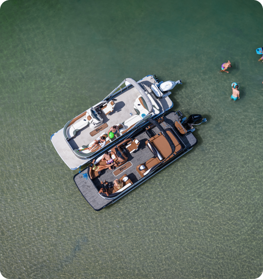 Aerial view of two connected motorboats in clear shallow water, with several people on board and a few others swimming nearby. The sunny, recreational scene highlights the perfect setting for enjoying time on the water, reminiscent of new pontoon boat sales adventures.
