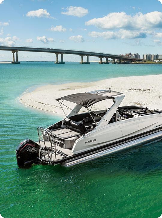 A sleek, modern boat with an outboard motor is anchored near a sandy strip of land in clear, turquoise water. The boat, equipped with a canopy, contrasts with the bright, sunny day under a blue sky with scattered clouds. In the background, a long bridge spans the water and hints at the cost of pontoon boats docked nearby.