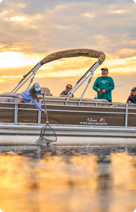 A group of people on a Sun Tracker pontoon boat are enjoying a fishing trip at sunset. One person leans over, using a net to catch a fish from the water, while others watch and smile. The sky is painted with warm sunset colors, reflecting the priceless moments shared regardless of the cost of a pontoon boat.