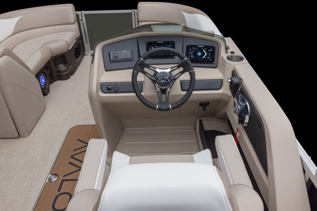 A steering area of a modern boat with tan upholstery. Features a steering wheel, digital dashboard, and control panel. The seating area is partially visible with beige cushions, and the boat flooring has a design element with the word "Avalon.