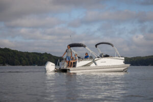 A silver pontoon boat with three people onboard is cruising on a calm lake. The sky is partly cloudy, and dense green trees line the distant shoreline. The group appears relaxed, enjoying the serene water setting.