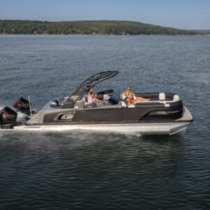 A sleek pontoon boat with multiple people aboard speeds across a calm body of water. The boat has a modern design and powerful engines, with a forested shoreline visible in the background under a clear blue sky.