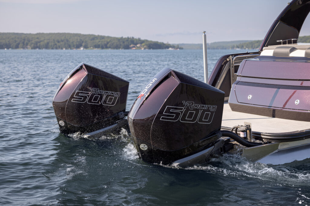 Two high-performance boat engines labeled "Racing 500" mounted on the back of a sleek boat, floating on a calm lake. The distant shoreline is visible under a clear sky.