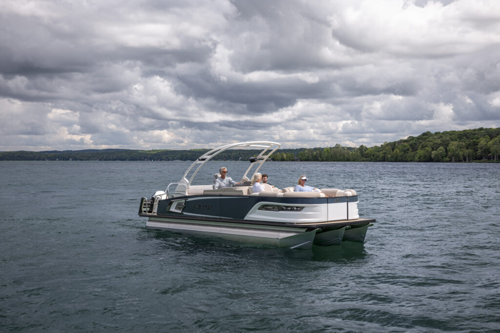 A pontoon boat with a canopy cruises on a lake under a cloudy sky. Four people onboard are sitting and enjoying the ride, surrounded by calm waters and a backdrop of green, forested shoreline.