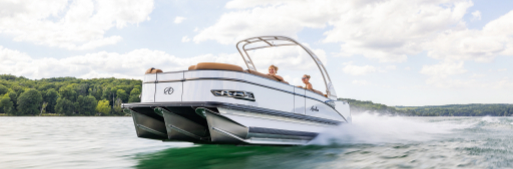 A sleek pontoon boat glides swiftly across a lake, creating a trail of white spray. Two people are seated inside, enjoying the sunny day. The shoreline with lush green trees is visible in the background under a partly cloudy sky.