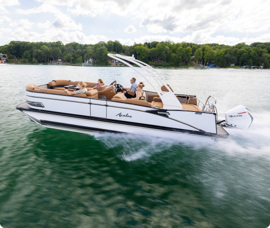 A sleek white pontoon boat with several passengers swiftly cruises across a lake surrounded by lush green trees under a partly cloudy sky. The boat creates ripples in the water as it moves forward.