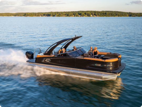 A sleek black pontoon boat speeds across a calm lake, carrying five passengers enjoying the ride. The background features a lush, green shoreline under a blue sky with scattered clouds.