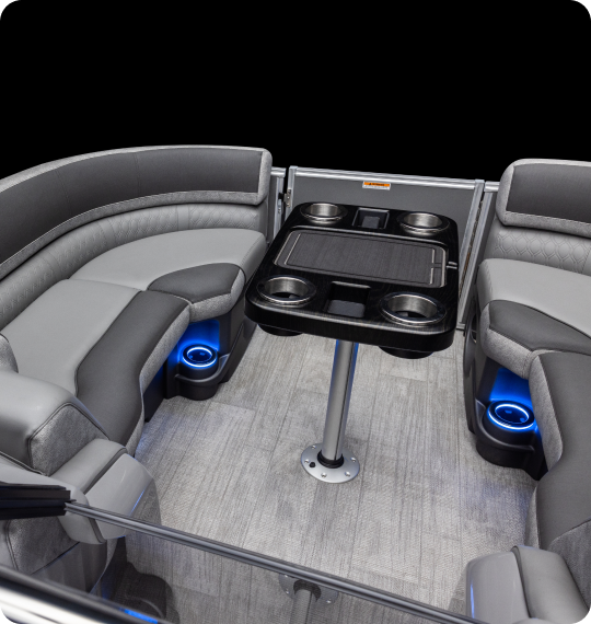 Interior of a luxury pontoon boat featuring curved gray seating with cup holders and blue LED lighting. A rectangular table with built-in cup holders is centered on a carpeted floor.