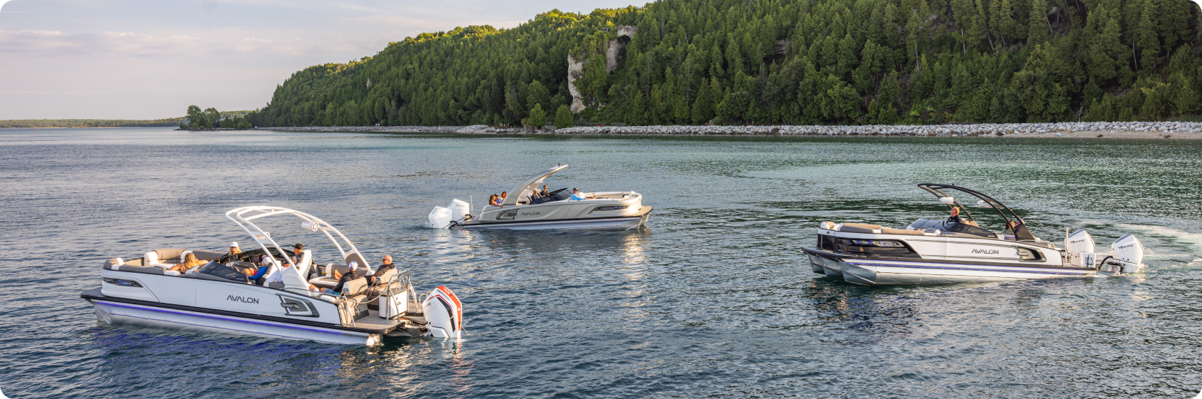 Three pontoon boats with people on board cruise on a calm lake, surrounded by dense green trees and a rocky shoreline under a partially cloudy sky.