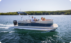 A family is enjoying a leisurely boat ride on a large lake surrounded by forested shores. The pontoon boat, with its canopy offering shade, glides across calm waters under the clear, sunny sky—a serene escape where thoughts of pontoon prices drift away with the gentle waves.