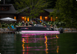 A sleek black boat with glowing pink lights reflects on a calm lake near a cozy lakeside cabin. People gather onshore, enjoying the tranquility as evening descends, contemplating pontoon prices for their next adventure in this serene setting surrounded by whispering trees.
