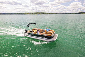A pontoon boat with several people on board travels across a large, calm lake under a partly cloudy sky, surrounded by distant tree-covered shoreline.