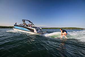 A person surfs on the water behind a motorboat with several people onboard, under a clear blue sky with a distant treeline in the background.