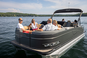 Five people relax and chat on a dark gray pontoon boat floating on a calm lake, surrounded by tree-lined shores under a sunny sky.
