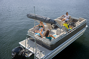 Four people relax on a pontoon boat floating on calm water. Two women sit on side benches, one man steers, and another woman lounges at the front. The boat has a canopy and visible outboard motor.