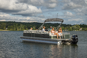 Four people sit and relax on a pontoon boat floating on a calm lake, surrounded by green trees and a partly cloudy sky.